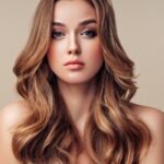 A close-up portrait of a young woman with long, light brown wavy hair, subtle layers framing her face, and natural makeup against a neutral beige background