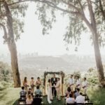 Outdoor micro wedding ceremony under tall trees with city view in the background.