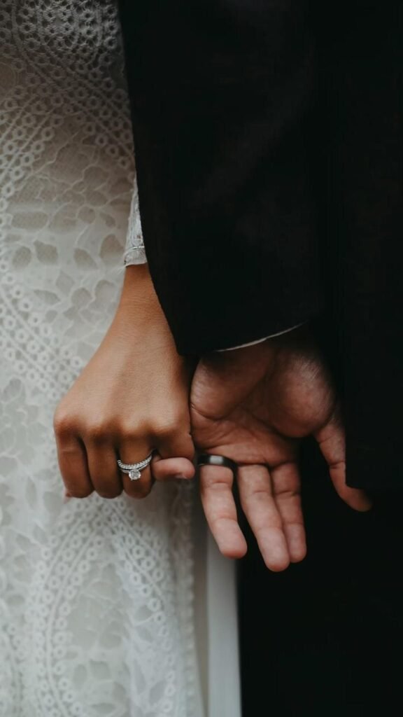 Close-up of bride and groom holding hands, showing wedding rings and lace bridal gown.