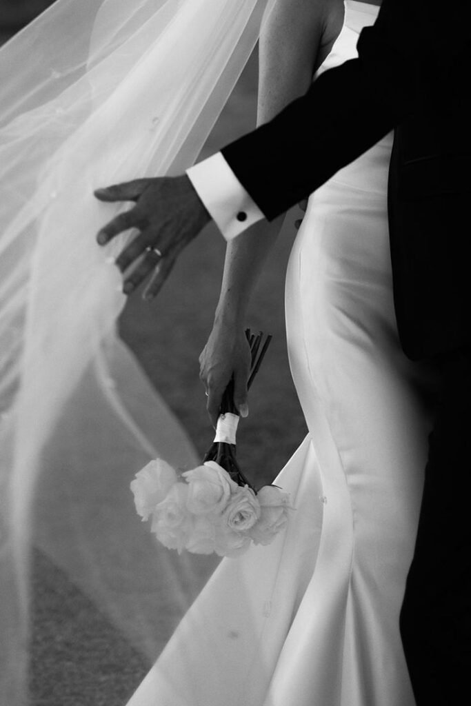 Bride and groom holding hands with white roses in elegant black and white wedding photo.