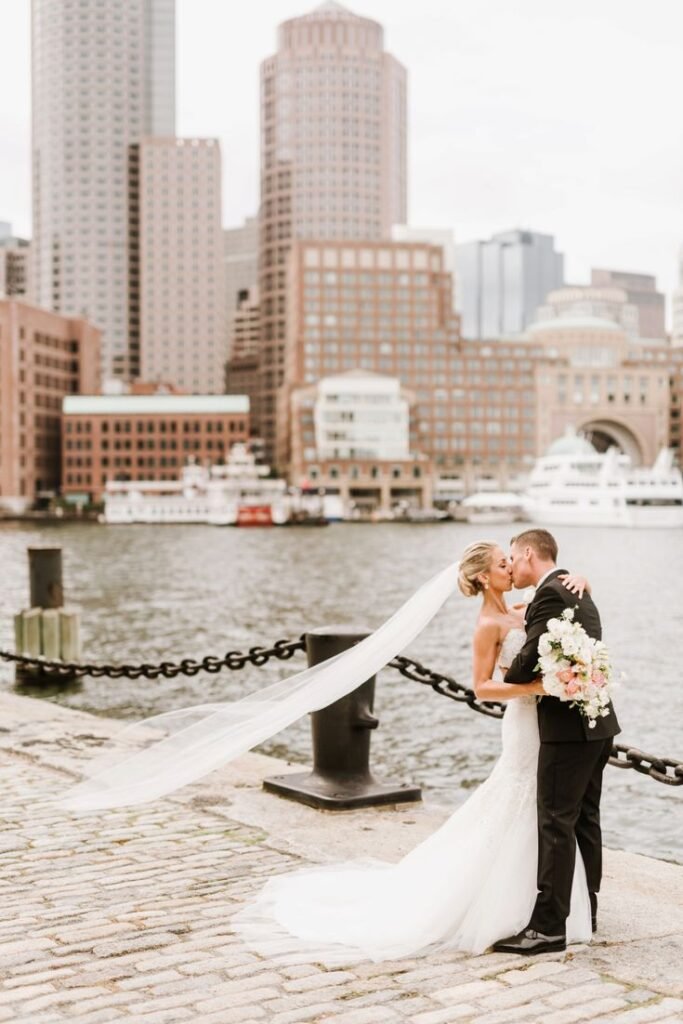 Bride and groom kissing by the Boston Harbor with skyline and waterfront view in the background.