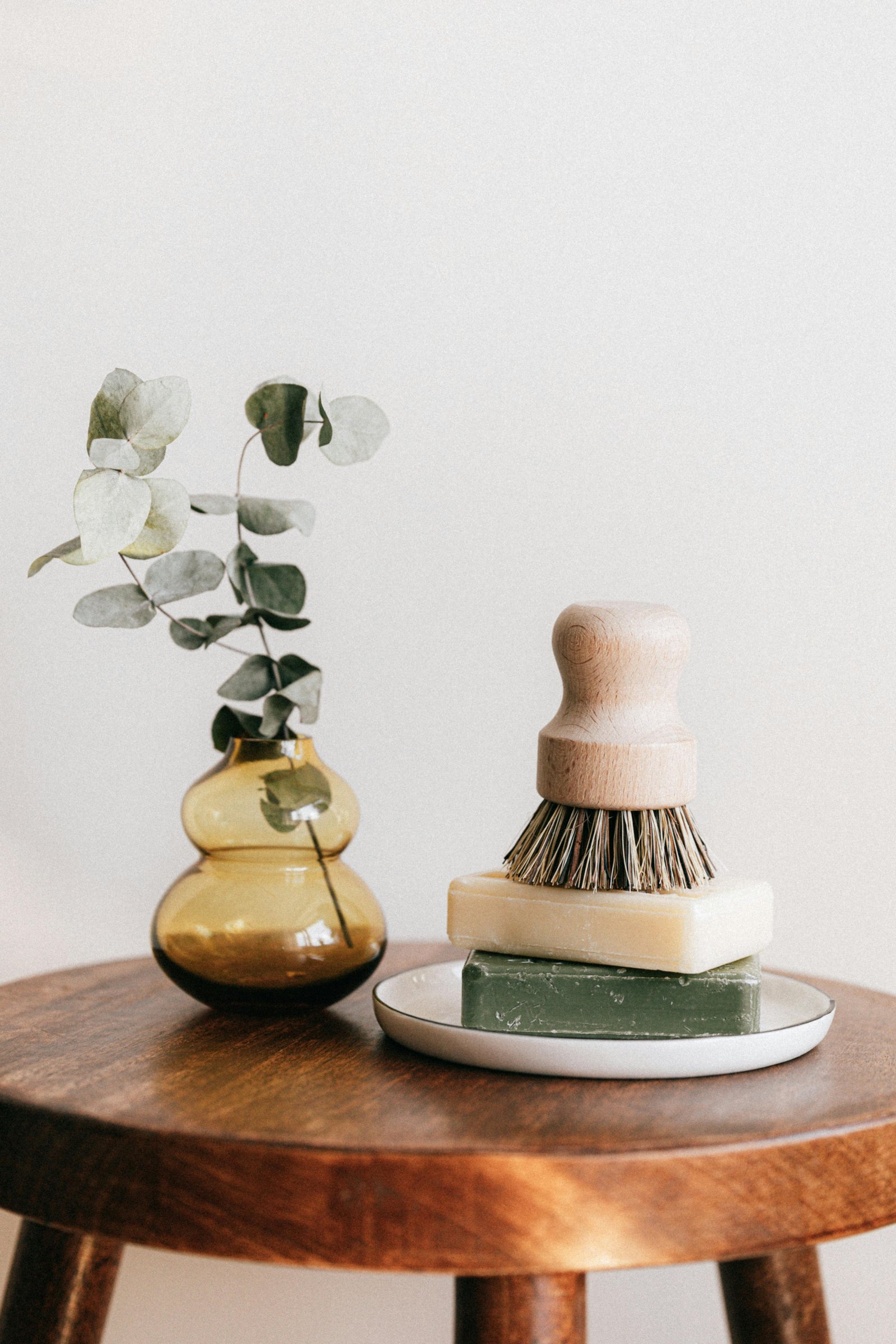 Minimalist spa decor with natural soap bars, a wooden brush, and eucalyptus leaves in an amber glass vase on a wooden stool.