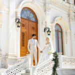 Bride and groom standing on elegant marble staircase of a grand wedding venue in Malibu, holding hands in front of ornate wooden doors.