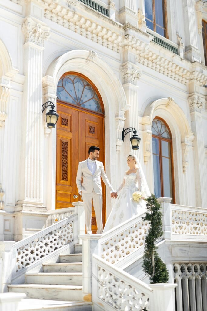 Bride and groom standing on elegant marble staircase of a grand wedding venue in Malibu, holding hands in front of ornate wooden doors.